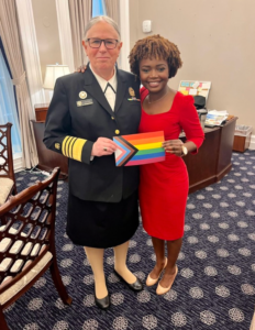 A person in uniform holding a rainbow flag and a person in a red dressDescription automatically generated