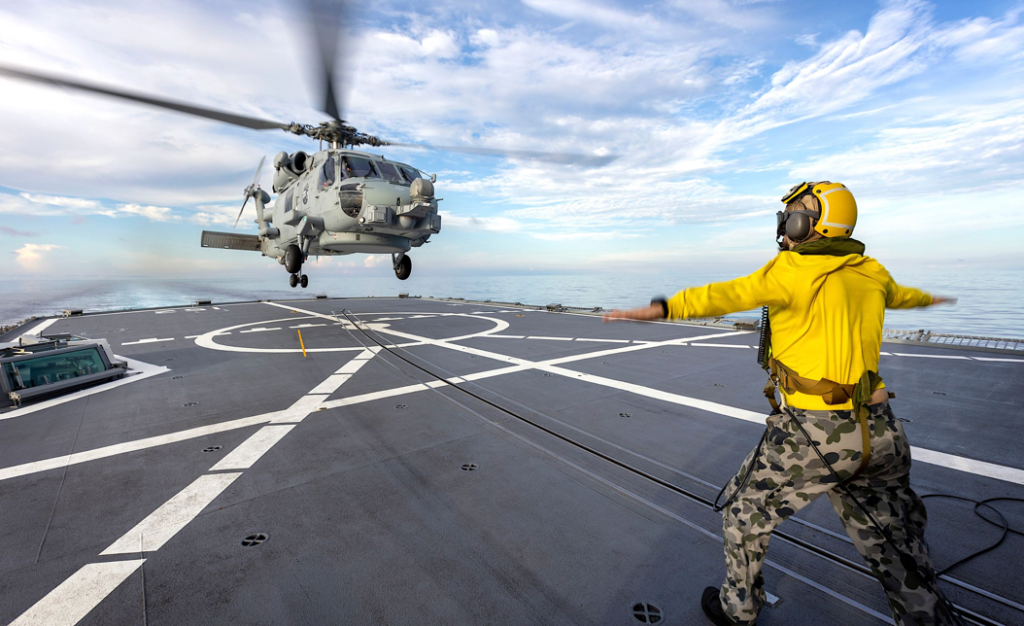 A person in a yellow shirt and camouflage pants standing on a helicopter landing padDescription automatically generated